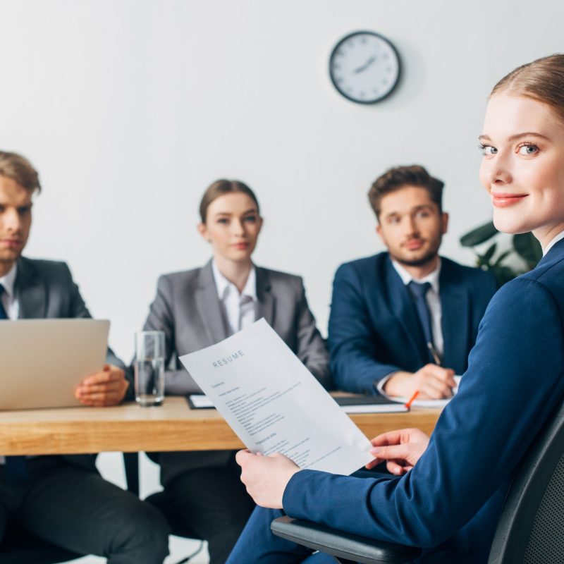 selective-focus-of-employee-looking-at-camera-while-holding-resume-near-recruiters.jpg